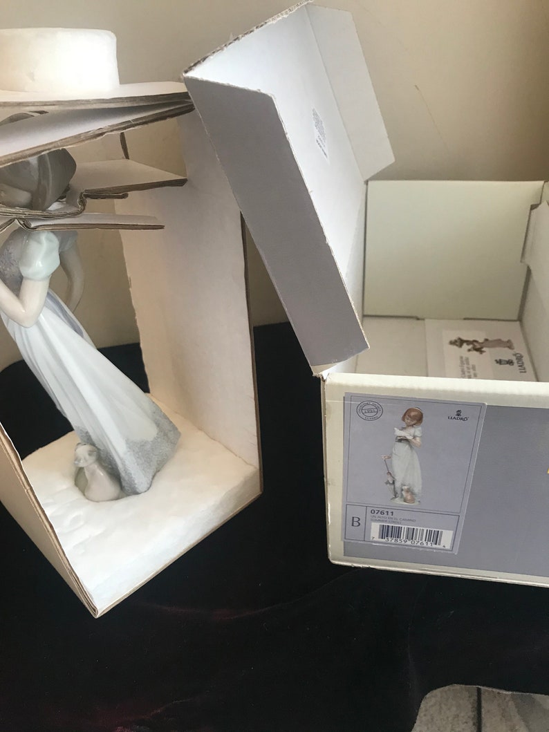 May include: A porcelain figurine of a girl in a white dress holding a book and a small white bird. The figurine is in its original box, which is white with a blue and gold label. The label includes the number 07611 and the brand name Lladro.