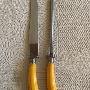 May include: A carving knife and sharpening steel set. The knife has a silver blade and a yellow handle. The sharpening steel is silver and has a matching yellow handle. The set is displayed on a textured, off-white surface.