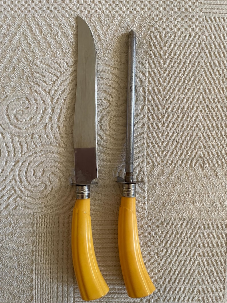 May include: A carving knife and sharpening steel set. The knife has a silver blade and a yellow handle. The sharpening steel is silver and has a matching yellow handle. The set is displayed on a textured, off-white surface.