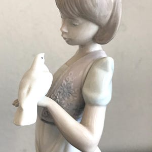 May include: A porcelain figurine of a girl with short brown hair holding a white dove. She is wearing a white dress with a floral vest.