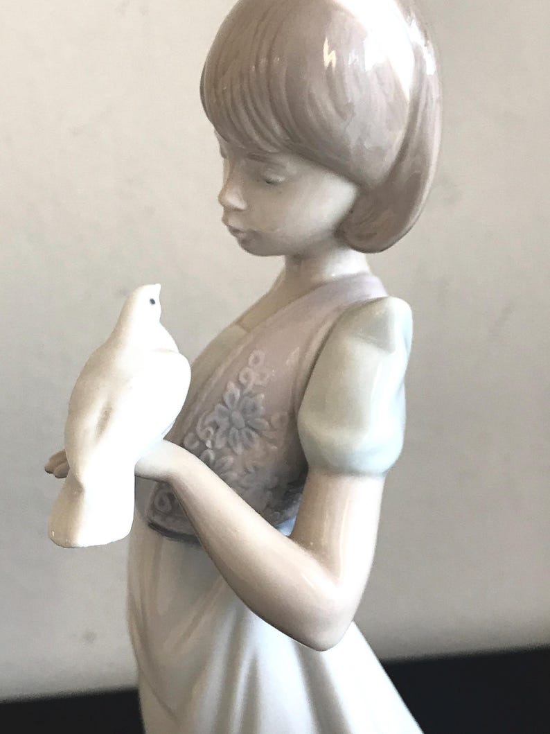 May include: A porcelain figurine of a girl with short brown hair holding a white dove. She is wearing a white dress with a floral vest.