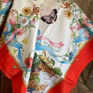 May include: A white silk scarf with a red border. The scarf features a floral design with butterflies, ribbons, and a basket of fruit. The basket has a label that says "Gucci".