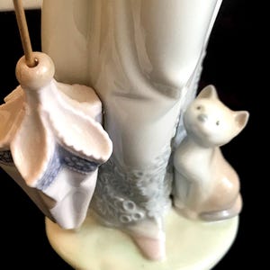 May include: A porcelain figurine of a woman holding an umbrella and a small cat. The woman is wearing a white dress with a floral pattern and the cat is sitting on the ground.
