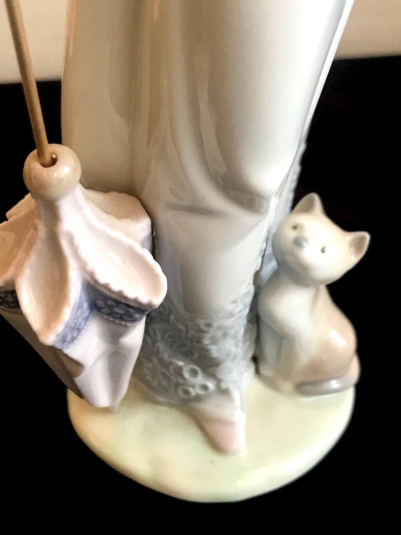 May include: A porcelain figurine of a woman holding an umbrella and a small cat. The woman is wearing a white dress with a floral pattern and the cat is sitting on the ground.
