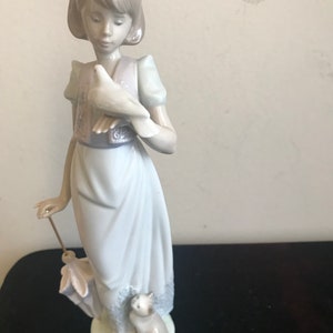 May include: A porcelain figurine of a girl holding a white dove and a small cat. She is wearing a blue dress with a pink vest and a white headband.