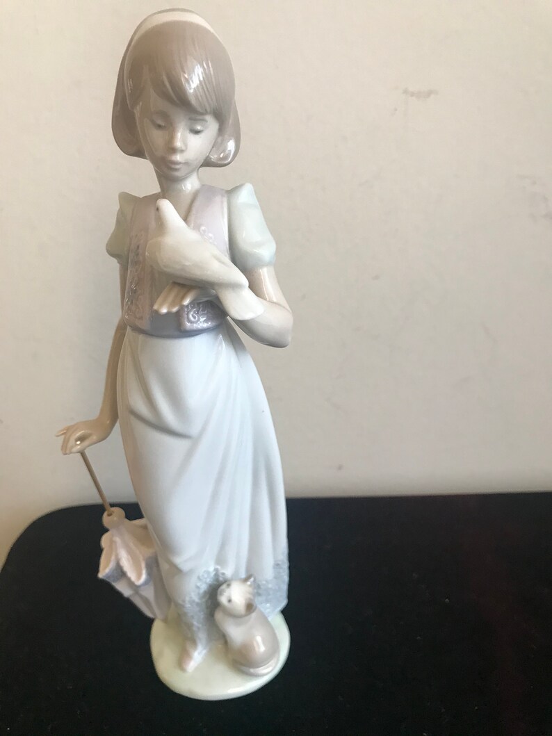 May include: A porcelain figurine of a girl holding a white dove and a small cat. She is wearing a blue dress with a pink vest and a white headband.