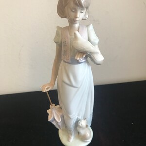 May include: A porcelain figurine of a girl holding a white dove in her right hand. She is wearing a light blue dress with a floral vest and a white headband. She is holding a closed umbrella in her left hand. A small white cat is sitting at her feet.