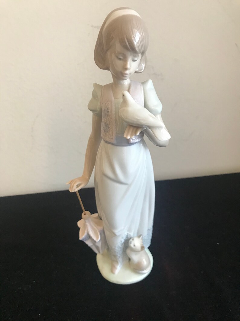 May include: A porcelain figurine of a girl holding a white dove in her right hand. She is wearing a light blue dress with a floral vest and a white headband. She is holding a closed umbrella in her left hand. A small white cat is sitting at her feet.