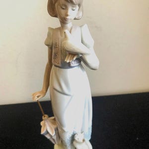 May include: A porcelain figurine of a girl holding a white dove in her right hand. She is wearing a light blue dress with a floral pattern and a brown vest. She is standing on a white base with a small white cat at her feet.