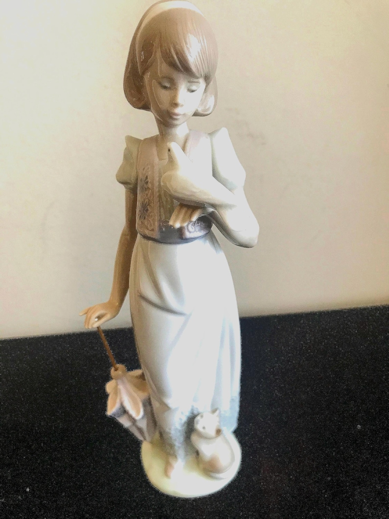 May include: A porcelain figurine of a girl holding a white dove in her right hand. She is wearing a light blue dress with a floral pattern and a brown vest. She is standing on a white base with a small white cat at her feet.