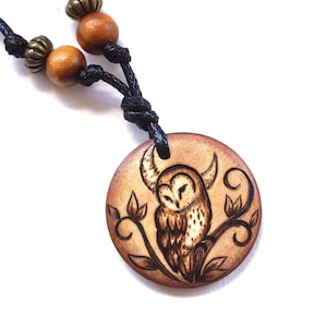 May include: A wooden pendant necklace with a pyrography owl design. The round pendant is attached to a black cord with wooden and metal beads. The owl is perched on a branch with leaves, all in shades of brown.