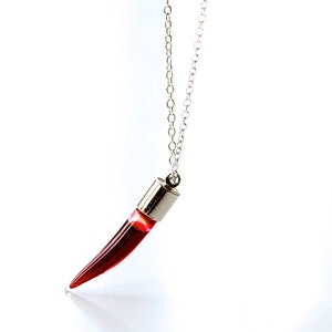 Vial necklace in the shape of a long fang or a chilli pepper.  Transparent glass vial with a silver lid. On a silver chain and filled with realistic fake blood.