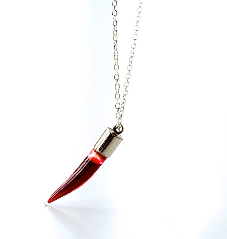 Vial necklace in the shape of a long fang or a chilli pepper.  Transparent glass vial with a silver lid. On a silver chain and filled with realistic fake blood.