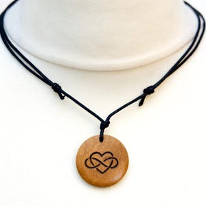 May include: A wooden necklace with a black cord and a round pendant featuring an infinity symbol inside a heart. The pendant is made of light brown wood.