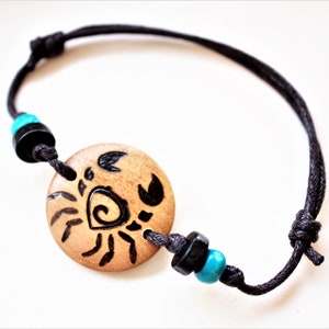 A bracelet with a wooden disc charm on an adjustable black cord with sliding knots.  The charm shows a wood burned design example of a stylized crab. Flanked with blue and black beads.