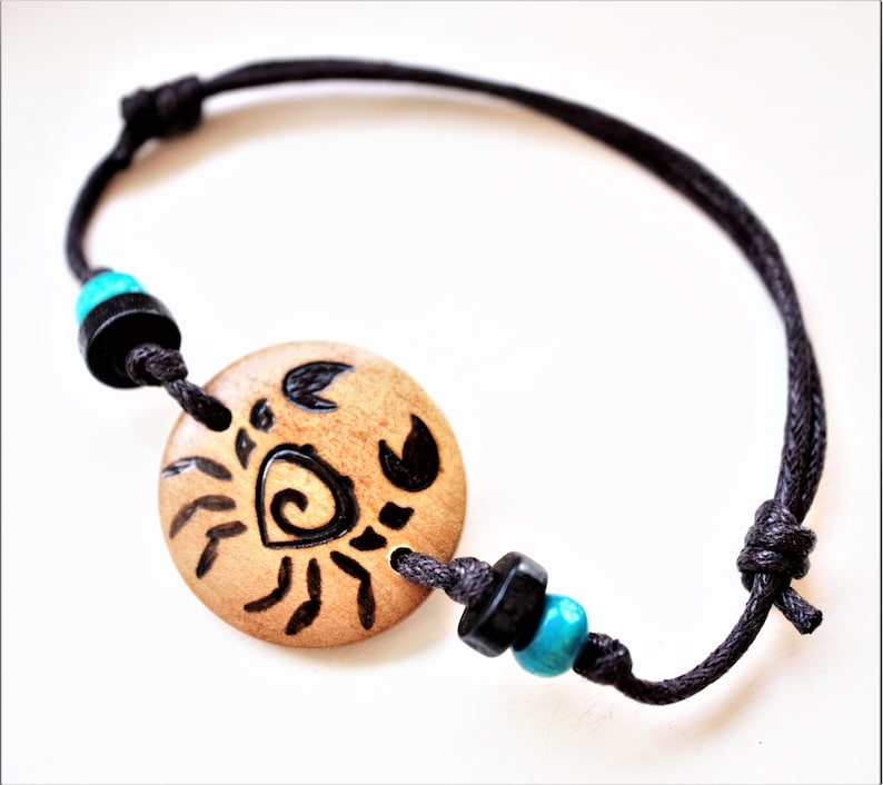 A bracelet with a wooden disc charm on an adjustable black cord with sliding knots.  The charm shows a wood burned design example of a stylized crab. Flanked with blue and black beads.