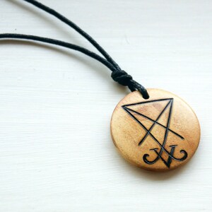 K&ouml;nnte beinhalten: Eine Holz-Halskette mit einem schwarzen Band. Der Anh&auml;nger ist rund und zeigt ein schwarz-wei&szlig;es Symbol eines Pentagramms mit einem Kreuz darin.