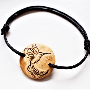 A bracelet with a wooden disc charm on an adjustable black cord with sliding knots.  The charm shows a wood burned design example of a stylized hummingbird.