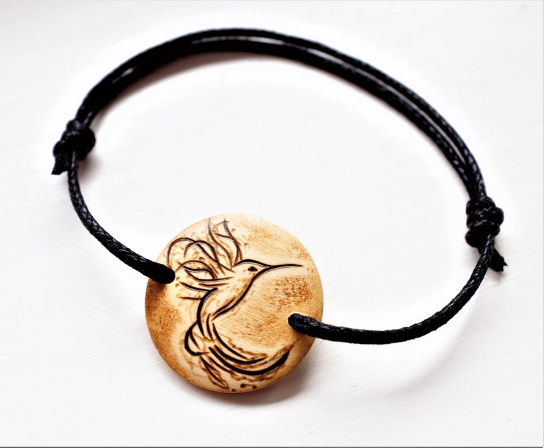 A bracelet with a wooden disc charm on an adjustable black cord with sliding knots.  The charm shows a wood burned design example of a stylized hummingbird.