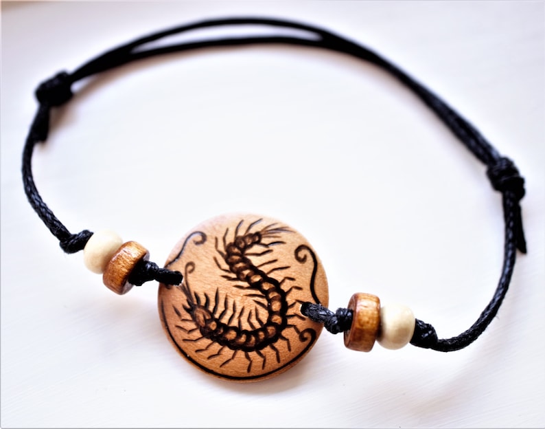 A bracelet with a wooden disc charm on an adjustable black cord with sliding knots.  The charm shows a wood burned design example of a centipede. Two small natural coloured wooden beads are threaded on to the cord at each side of the charm.