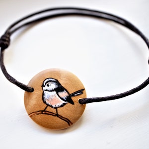 A bracelet with a wooden disc charm on an adjustable black cord with sliding knots.  The charm shows a wood burned design example of a longtailed tit.  The tiny bird is painted by hand.