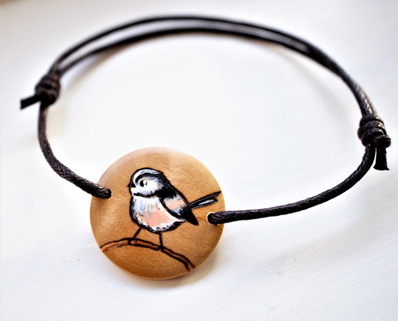 A bracelet with a wooden disc charm on an adjustable black cord with sliding knots.  The charm shows a wood burned design example of a longtailed tit.  The tiny bird is painted by hand.