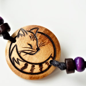 A bracelet with a wooden disc charm on an adjustable black cord with sliding knots.  The charm shows a wood burned design example of a sleeping cat. A purple and brown bead sit to each side of the charm.
