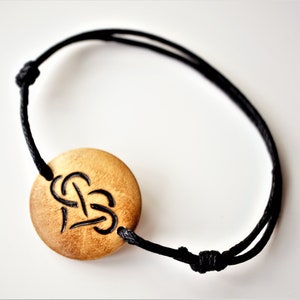 A bracelet with a wooden disc charm on an adjustable black cord with sliding knots.  The charm shows a wood burned design example of a heart, with a infinity symbol tied through the middle.