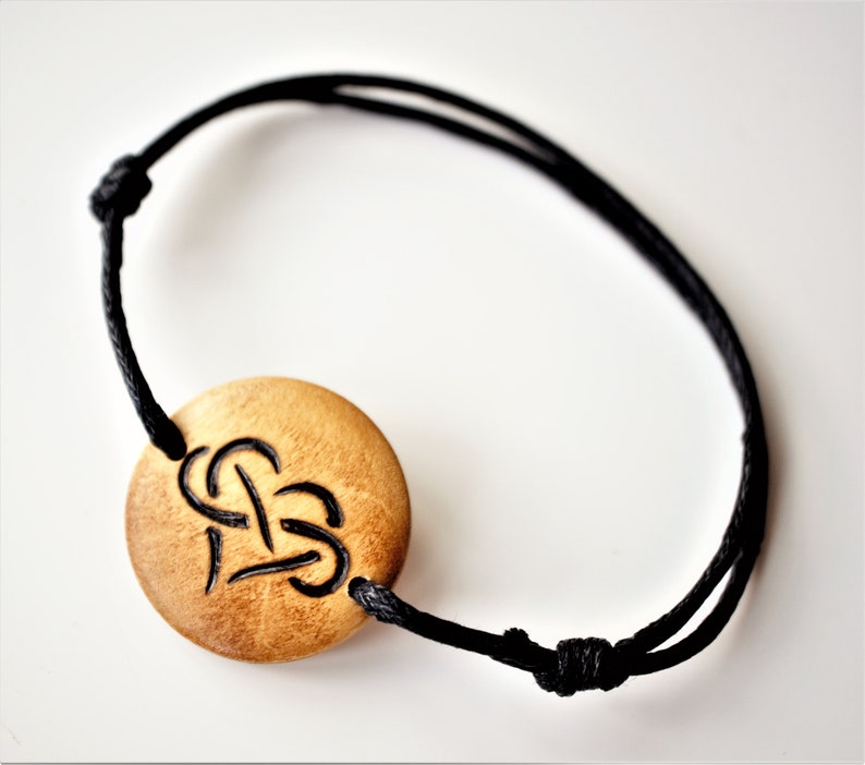 A bracelet with a wooden disc charm on an adjustable black cord with sliding knots.  The charm shows a wood burned design example of a heart, with a infinity symbol tied through the middle.