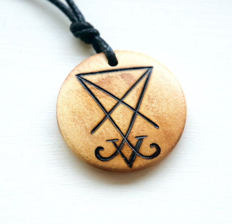 K&ouml;nnte beinhalten: Ein Holzanh&auml;nger mit einer schwarzen Schnur, der ein schwarzes Sigil mit den Buchstaben "V" und "V" zeigt, die in ein Dreieck eingeschrieben sind.