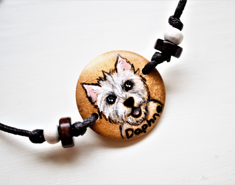 A bracelet with a wooden disc charm on an adjustable black cord with sliding knots.  The charm shows a wood burned design example of a pet dog, a highland terrier, persoanlised with the pets name underneath.