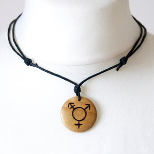 Puede incluir: Un collar con colgante de madera con un colgante redondo de color marrón claro con un símbolo transgénero negro. El colgante cuelga de un collar de cordón negro. El collar se muestra en un maniquí blanco.