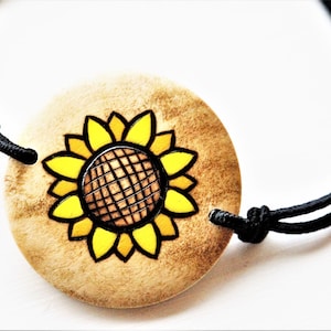 A bracelet with a wooden disc charm on an adjustable black cord with sliding knots.  The charm shows a wood burned design example of a sunflower.  The design is painted with shades of brigh yellow.