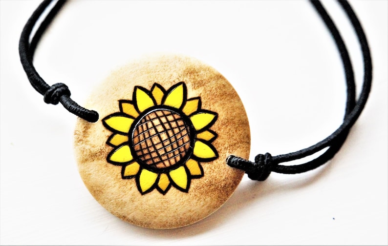 A bracelet with a wooden disc charm on an adjustable black cord with sliding knots.  The charm shows a wood burned design example of a sunflower.  The design is painted with shades of brigh yellow.