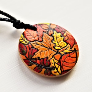 Pode incluir: Um colar com um pingente redondo de cerâmica com um design de folhas de outono coloridas. O pingente apresenta folhas vermelhas, laranja, amarelas e verdes.