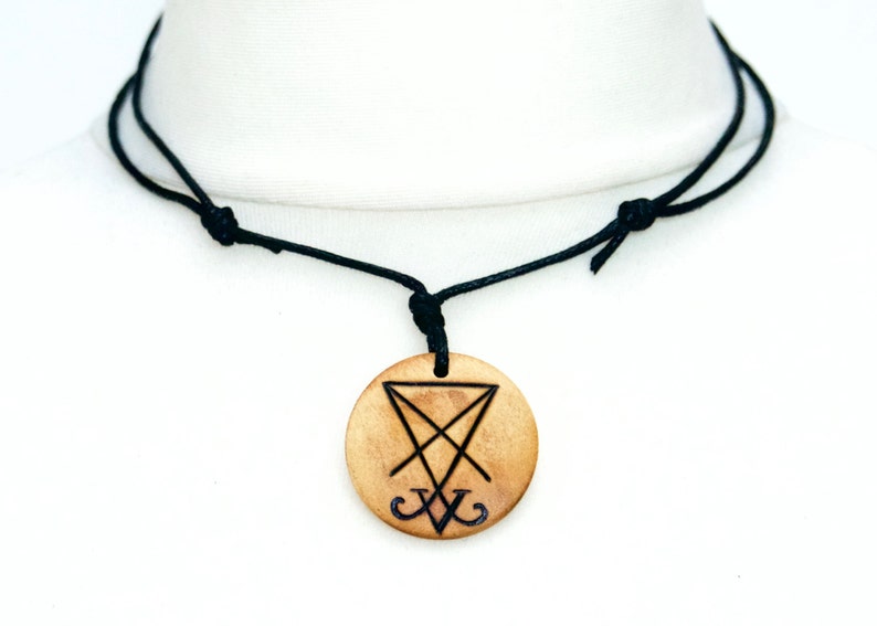 K&ouml;nnte beinhalten: Eine schwarze Schnurkette mit einem Holzanh&auml;nger mit einem schwarzen Sigil. Der Anh&auml;nger ist rund und hat eine hellbraune Farbe.