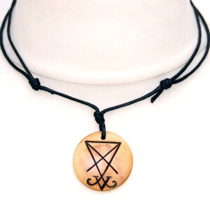 K&ouml;nnte beinhalten: Eine schwarze Schnurkette mit einem runden Holzanh&auml;nger mit einem schwarzen Sigil. Der Anh&auml;nger ist mit einem Dreieck und einem Kreuz verziert.