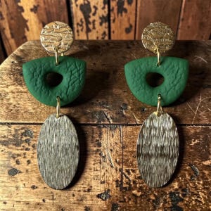 May include: A pair of handmade earrings with a gold-colored textured disc, a green, textured, half-moon shape with a hole, and a gold-colored oval pendant. The earrings are hanging on a wooden surface.