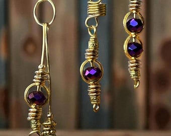 May include: Gold-toned metal hair jewelry with purple beads. The set includes three pieces: two single pieces and one longer piece with a cluster of purple beads at the bottom. The pieces have intricate spiral and bead detailing.