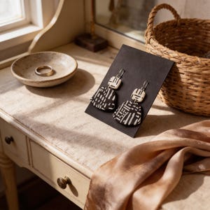 a pair of black and white patterned earrings displayed on a black card, placed on a wooden table next to a woven basket.