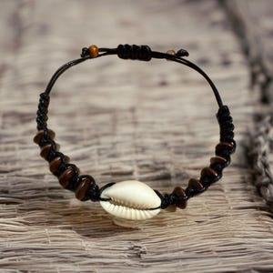 May include: A handmade bracelet featuring a white seashell centerpiece, brown beads, and a black woven cord. The adjustable bracelet is approximately 7 inches in diameter.