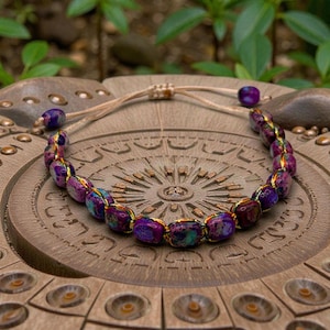 May include: A beaded bracelet featuring purple, blue, and gold-colored beads. The bracelet is strung on a beige cord with adjustable ends. The beads have a marbled appearance. The bracelet is displayed on a wooden surface.