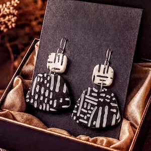 A pair of black and white patterned earrings are displayed in a black box with a gold lining, placed on a wooden surface.
