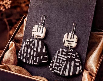 May include: A pair of dangle earrings featuring a small square top piece and a larger trapezoidal bottom piece, both in black with white etched line patterns. The earrings are attached to a black display card inside a box with gold lining.