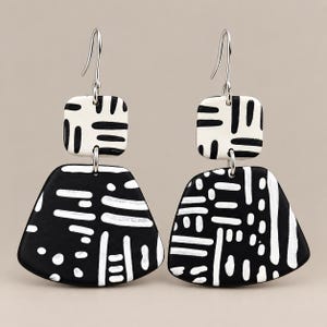 May include: A pair of dangle earrings featuring a small square top piece and a larger trapezoidal bottom piece. Both components are black with white hand-painted dash and dot patterns. The earrings hang from silver-toned metal ear wires.