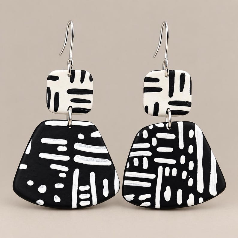 May include: A pair of dangle earrings featuring a small square top piece and a larger trapezoidal bottom piece. Both components are black with white hand-painted dash and dot patterns. The earrings hang from silver-toned metal ear wires.