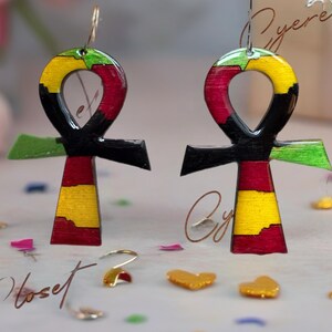 May include: A pair of Ankh-shaped earrings with a colorful design. The earrings feature a combination of black, red, yellow, and green. The earrings are suspended from silver-colored hooks. The background is a soft, blurred mix of colors.
