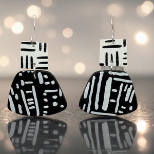 May include: A pair of black and white geometric earrings. Each earring has a square top with a linear design and a larger, teardrop-shaped bottom with a contrasting pattern. The earrings are suspended from silver-coloured hooks.