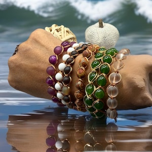 May include: A collection of beaded bracelets in various colors, including purple, white, brown, green, and clear. The bracelets are made with gold-colored wire and are displayed against a backdrop of ocean waves. The bracelets are a stylish accessory.