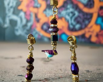 May include: Three gold-toned hair accessories with decorative beads and charms. One features a small bird charm with purple and blue details. The other two have dark purple and clear crystal beads. The accessories are designed to be inserted into the hair.